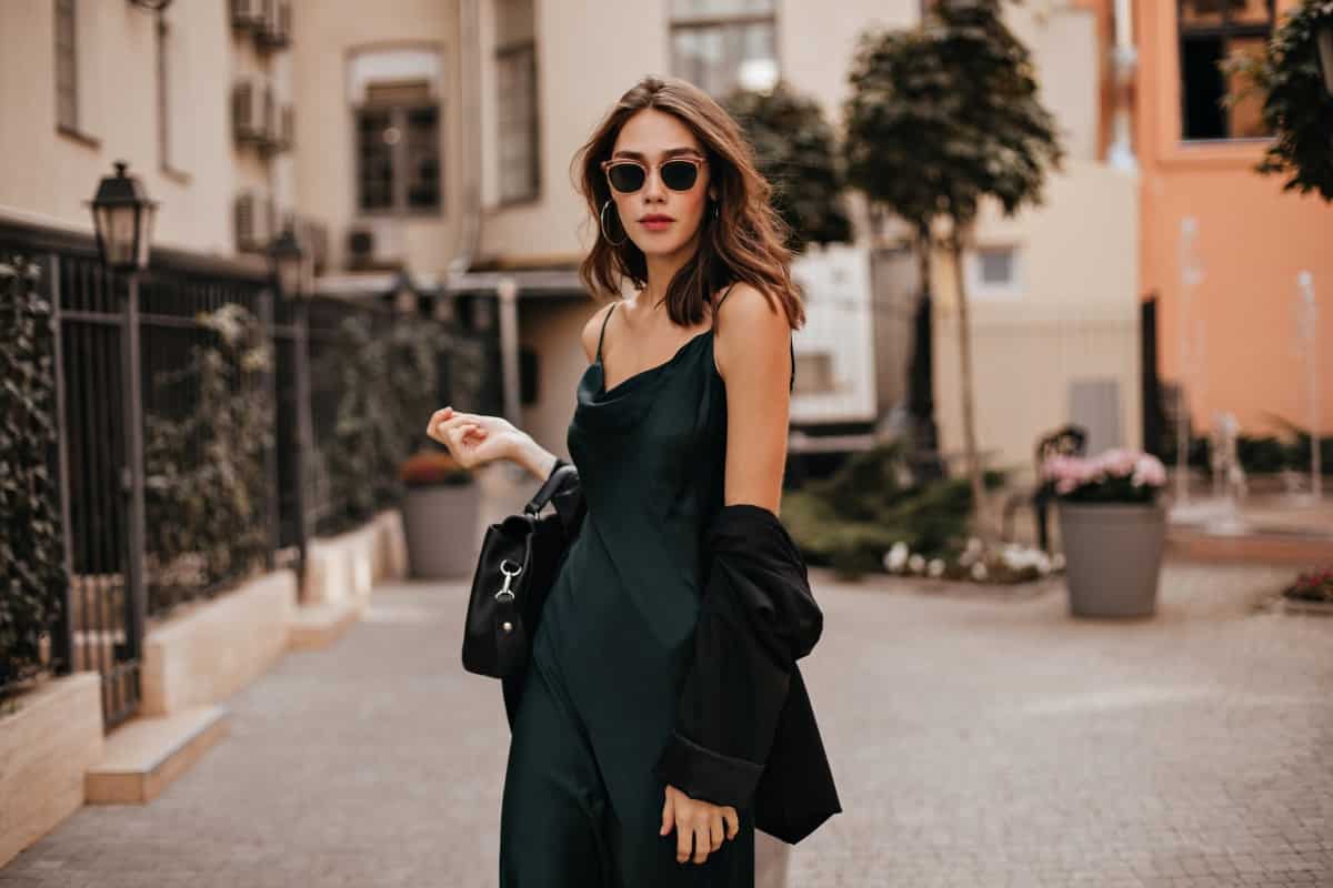 fashionable-pale-brunette-long-green-dress-black-jacket-sunglasses-standing-street-during-daytime-against-wall-light-city-building-1.jpg